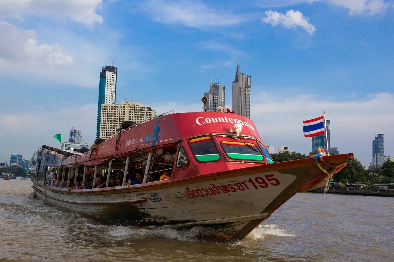 A wooden express boat with a coloured flag on its bow drawing alongside a pier on the Chao Phraya River