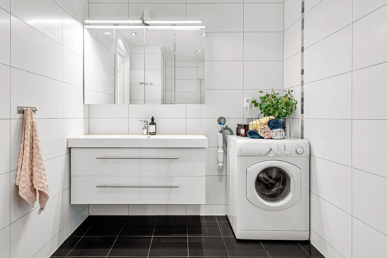 A washing machine mid-cycle in a small bathroom with folded laundry on a stool beside it