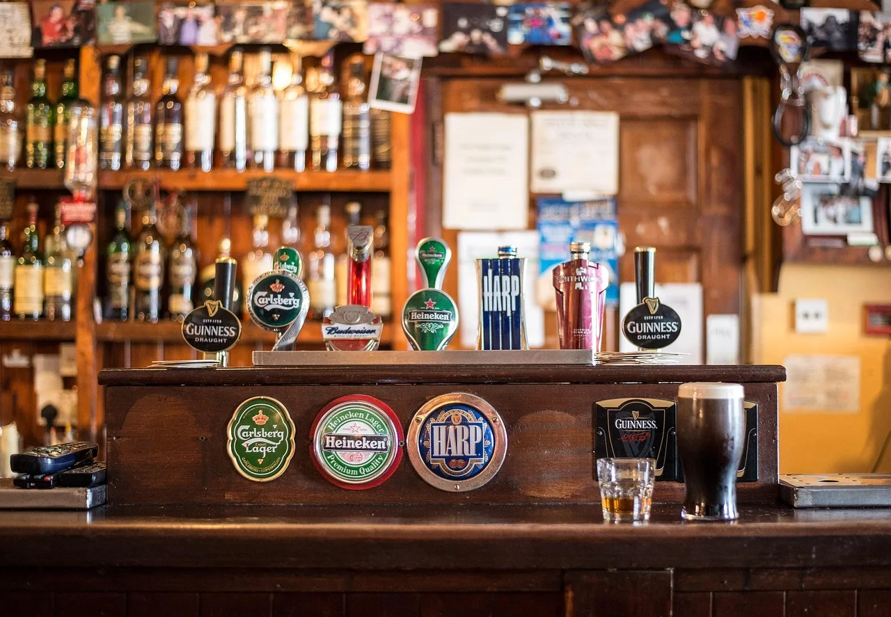 A row of hand pumps along a wooden pub bar counter with people standing loosely alongside