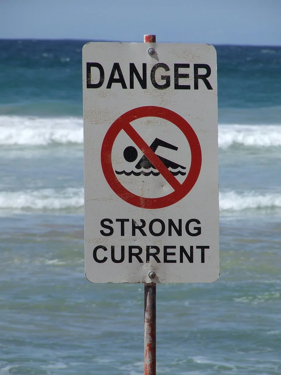 A red warning flag planted in wet sand at the water's edge with surf breaking behind