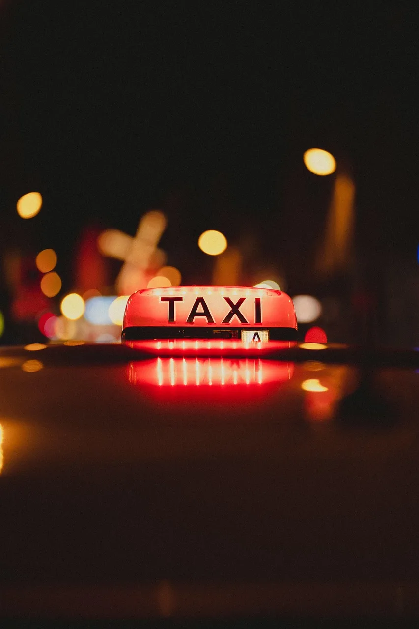 Passengers queueing at a signposted airport taxi rank while other drivers wait near the exit