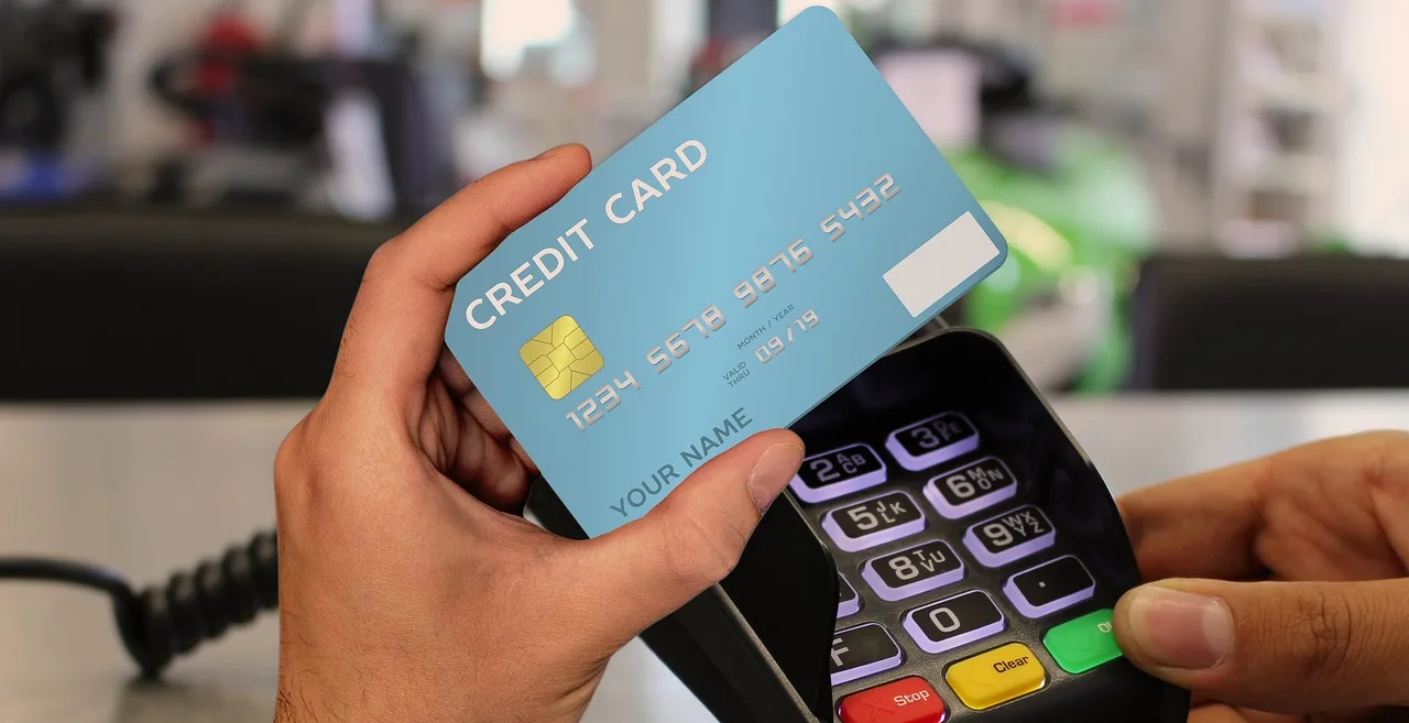 A hand holding a card above a payment terminal whose screen shows two currency amounts