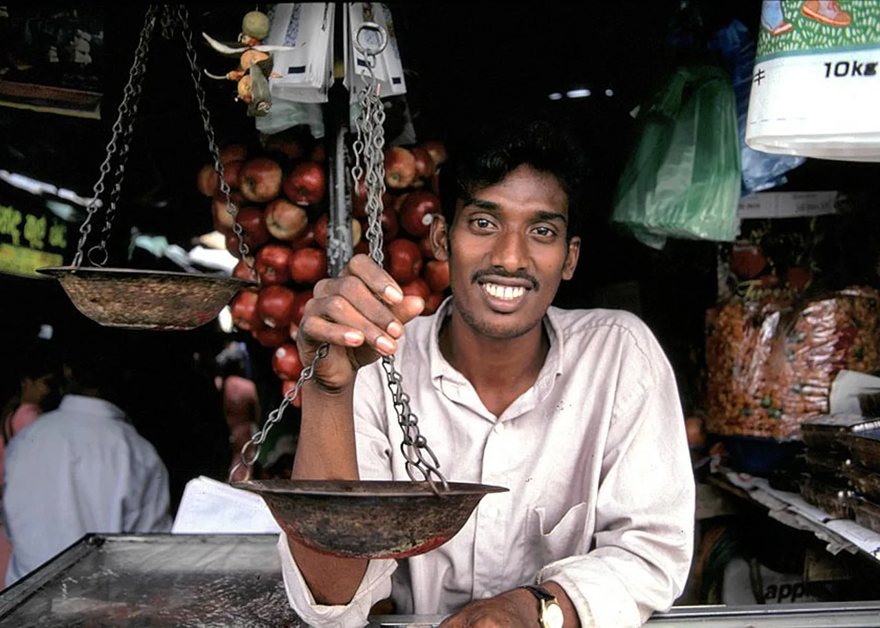 A covered bazaar lane lined with brass trays and hanging lanterns, a shopkeeper talking with a customer